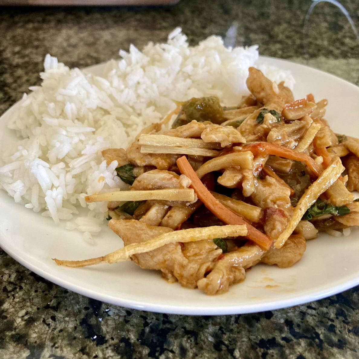 A plate on a counter is half full with rice and half full with a stir fry of chicken, carrots, chilies, and bamboo shoots.