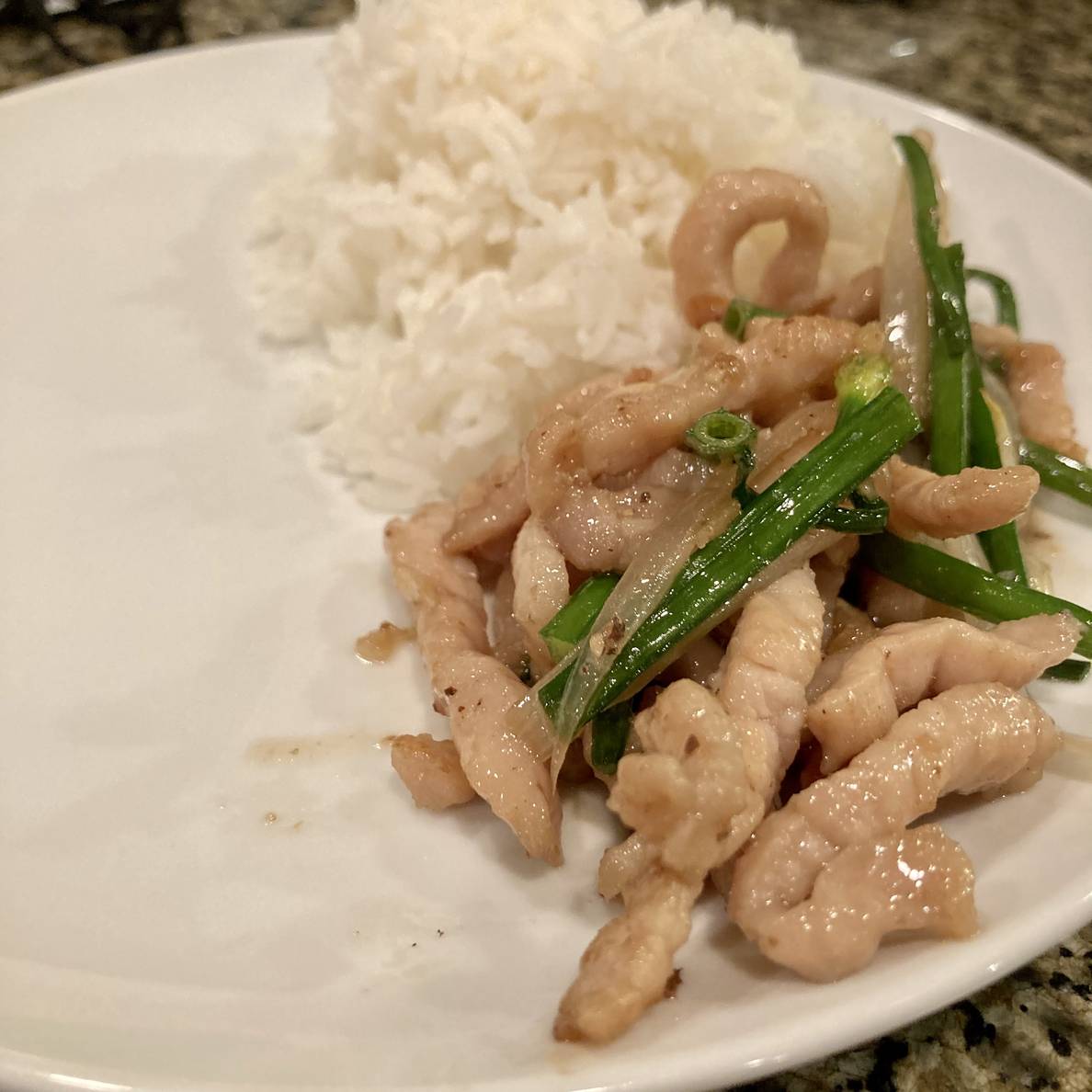 A plate on a counter has some rice out of focus in the background and some stir-fried pork with chives in focus in the foreground.