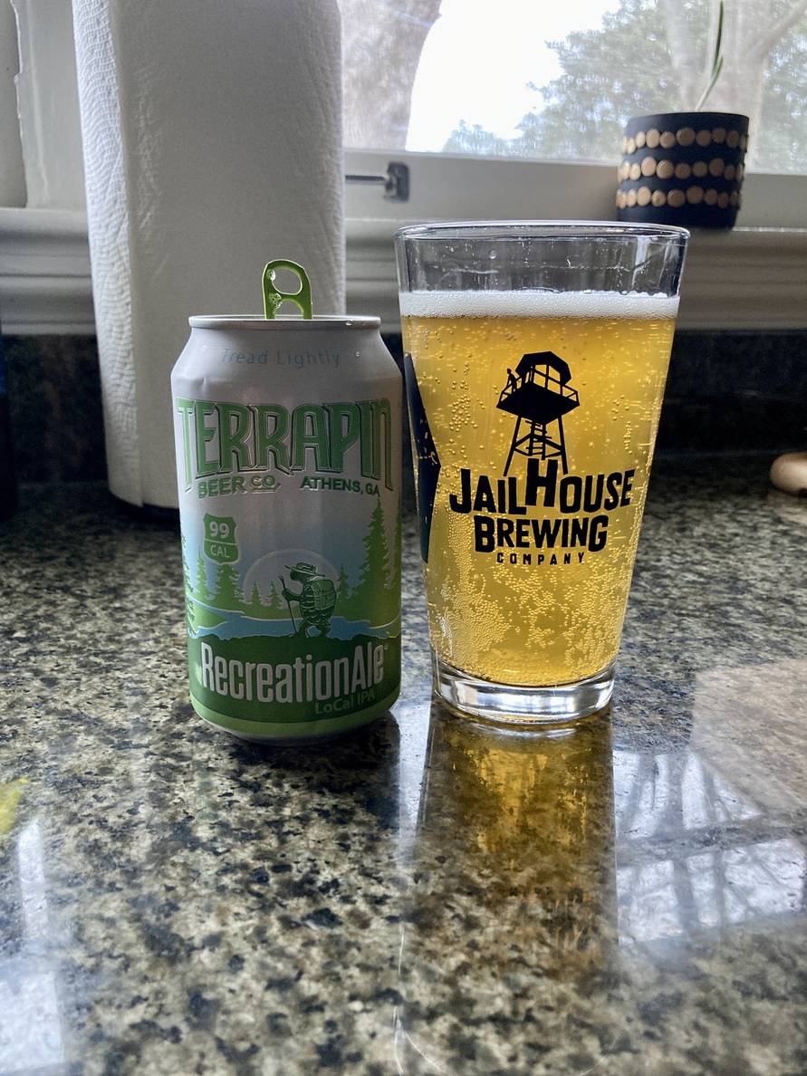 A beer can and beer glass sit on a counter. The can says Terrapin Recreation Ale. The glass is full of beer with sunlight coming from behind making it look bright. The glass says JailHouse Brewing Company.