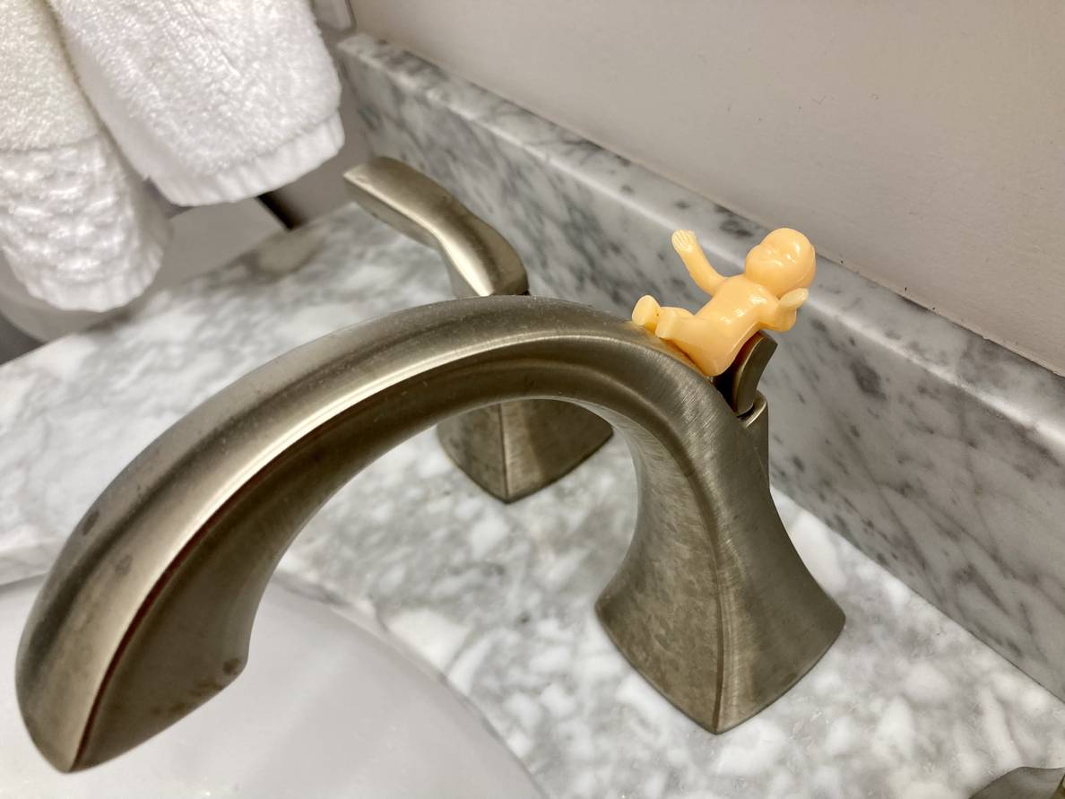 A small plastic baby rests delicately balanced on a bathroom sink faucet.