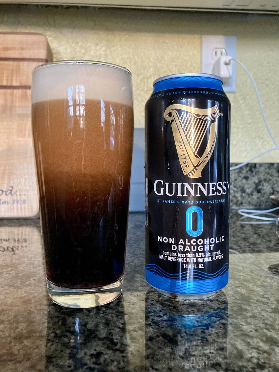 A beer glass with Guinness non-alcoholic beer cascading sits in a counter next to the can which Guinness 0 Non-Alcoholic Draught.