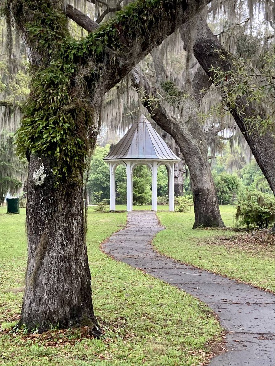 A concrete path winds from the foreground to a gazebo. Live oaks flank the path framing the gazebo. Spanish moss hangs from the branches.