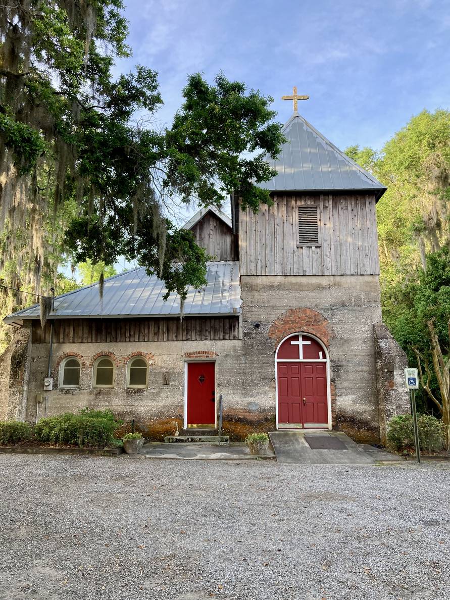 An old church with red doors made with a combination of masonry and wood, all weathered. A cross tops a two story massing on the right. A live oak frames the picture on the left.