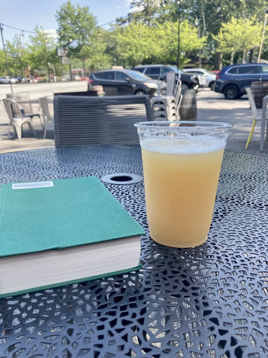 A plastic cup with hazy golden beer sits outside on a table alongside a book.