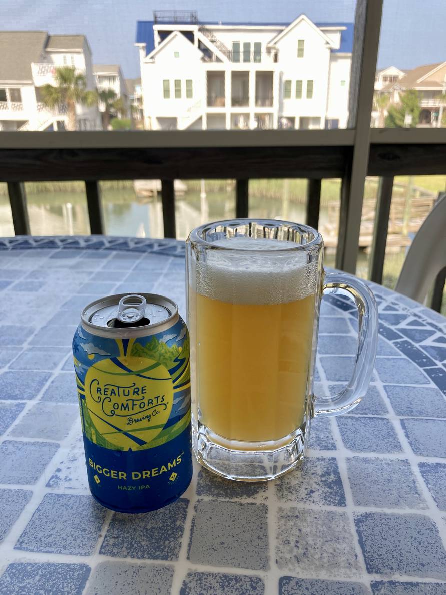 A beer can and a beer mug full of beer sit on an outdoor table. The can says ‘Creature Comforts Brewing Co. Bigger Dreams Hazy IPA’. In the background are beach houses and piers on a canal.