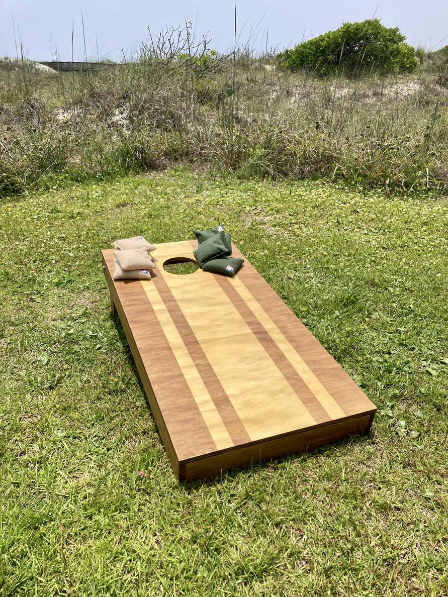 A cornhile board with natural wood stained in a vertical stripe pattern sits on a patch of grass. Hunter green and khaki bags sit on top of it.