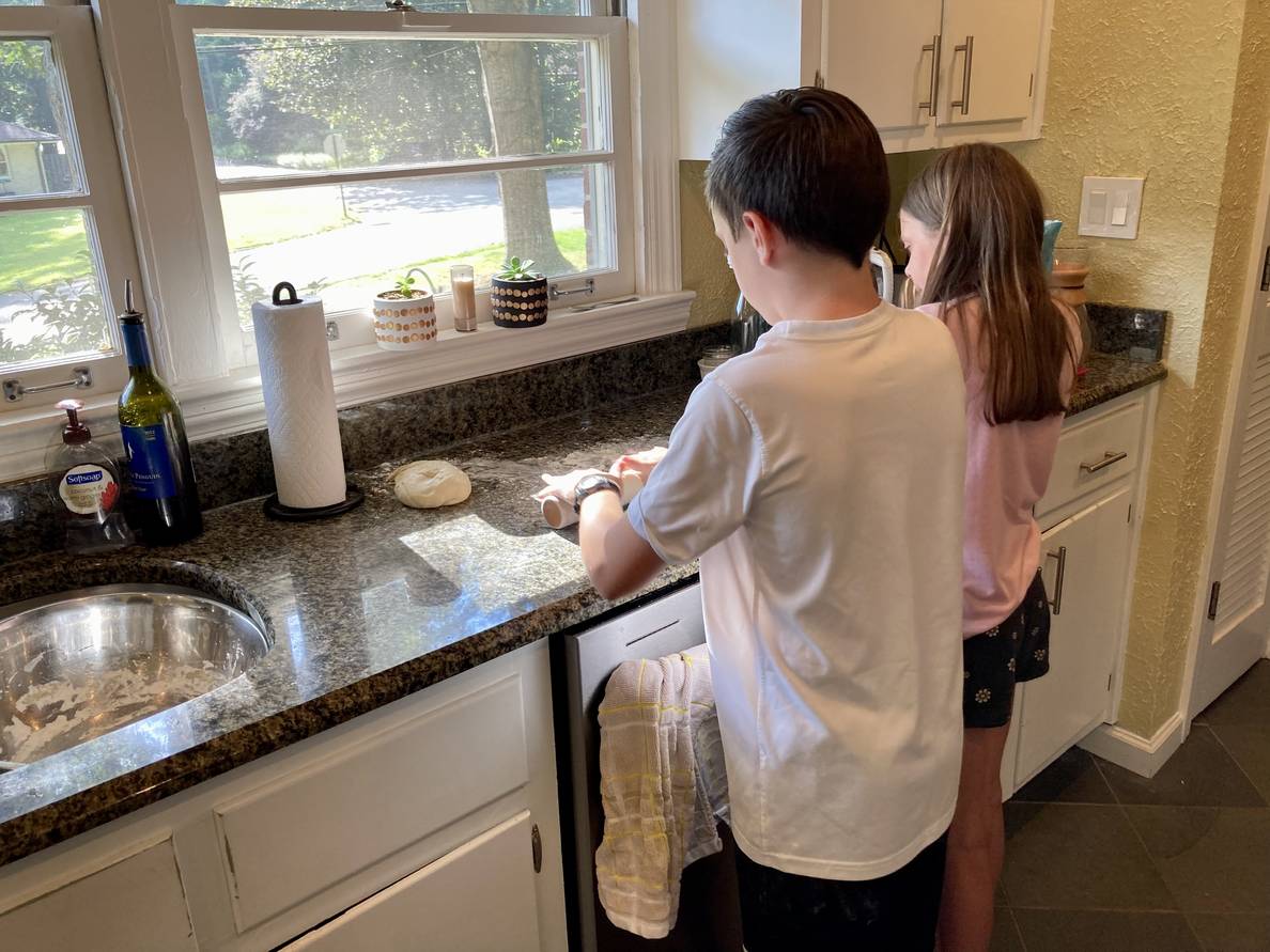 Two children stand in a kitchen, backs to the camera. They are working with a dough on the counter.