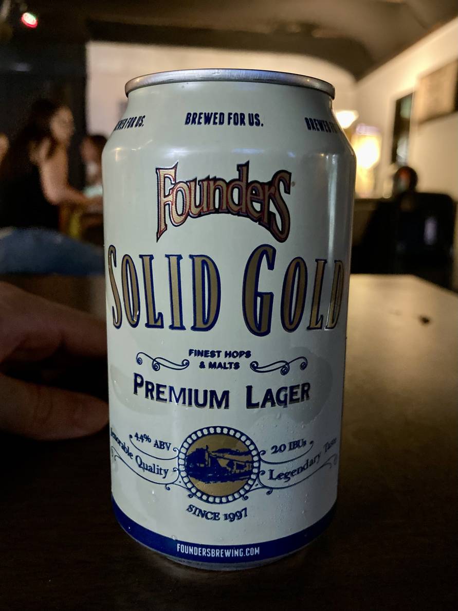 A white beer can sits on a table in a dim room. The can reads “Founder’s Solid Gold Premium Lager.”