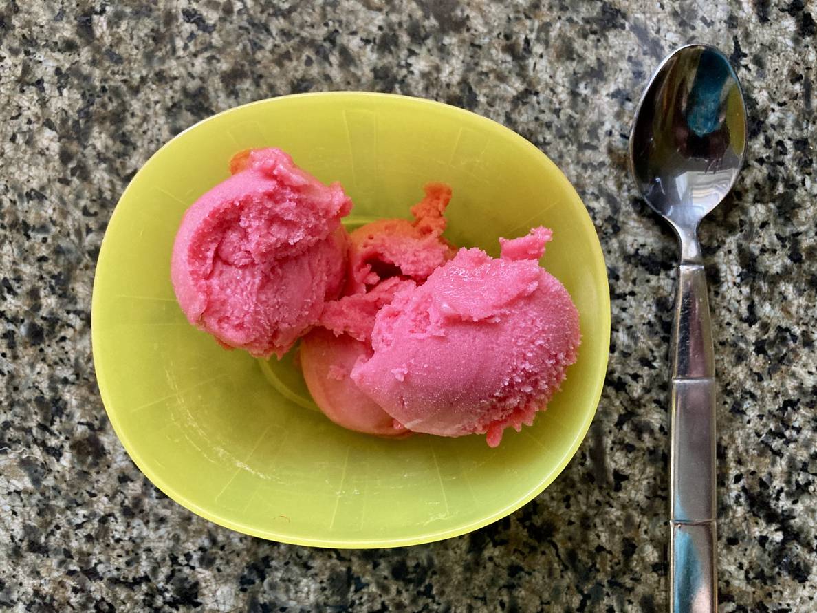 Three scoops of pink sorbet in a yellow plastic bowl on a granite countertop. A metal spoon rests next to the bowl.