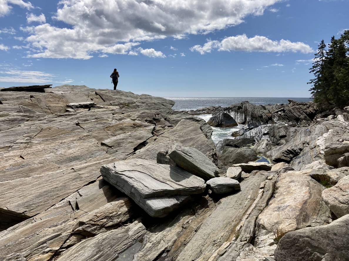 A woman in the distance stands atop rocks. More rocks extend from the foreground to ocean and rocky shore in the background.