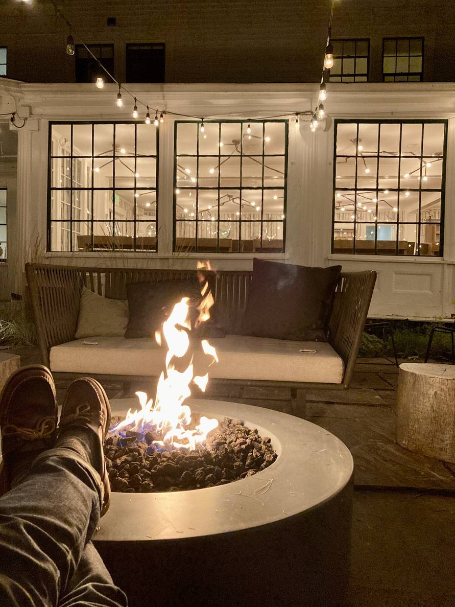 A fire put surrounded by chairs. A building with lighting and windows in the background. The photographer’s legs extend from the frame to rest on the fire pit ledge.