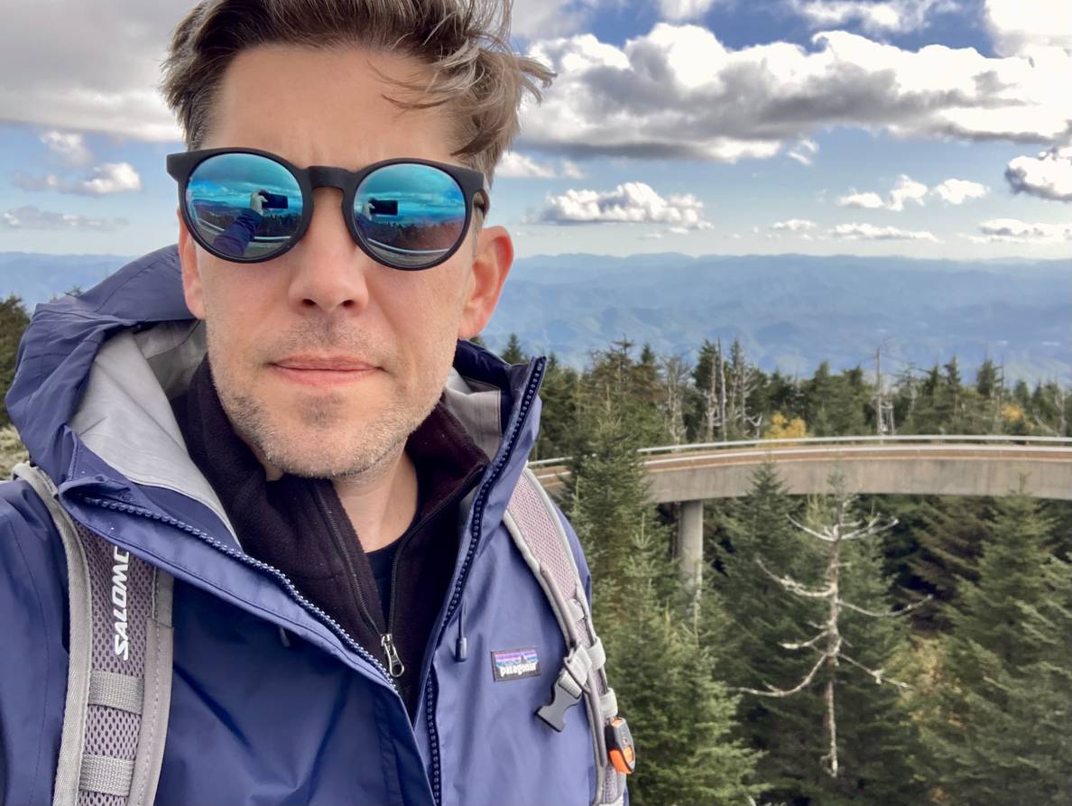 A selfie of a man with sunglasses, a backpack, and a rain jacket. Behind him a concrete ramp emerges from evergreen trees. Behind that ridges of mountains fade into the distance.