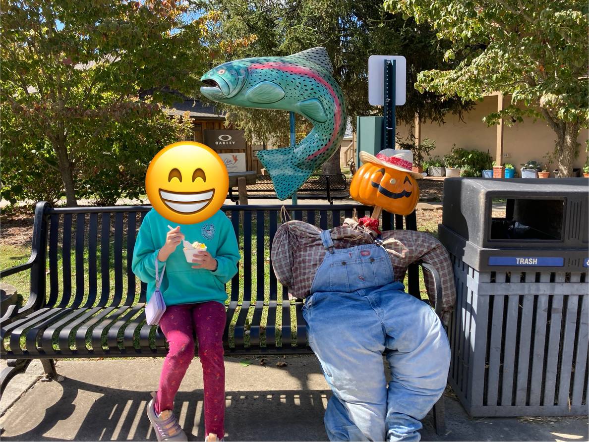A girl sits on a bench with a pumpkin-headed scarecrow. Behind them is a trout sculpture.