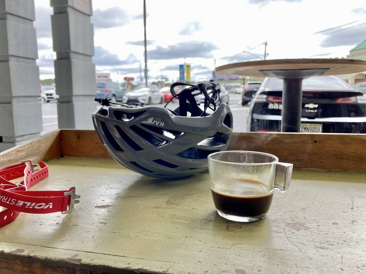 A glass of espresso on a wooden counter outside. A bike helmet is behind the glass.