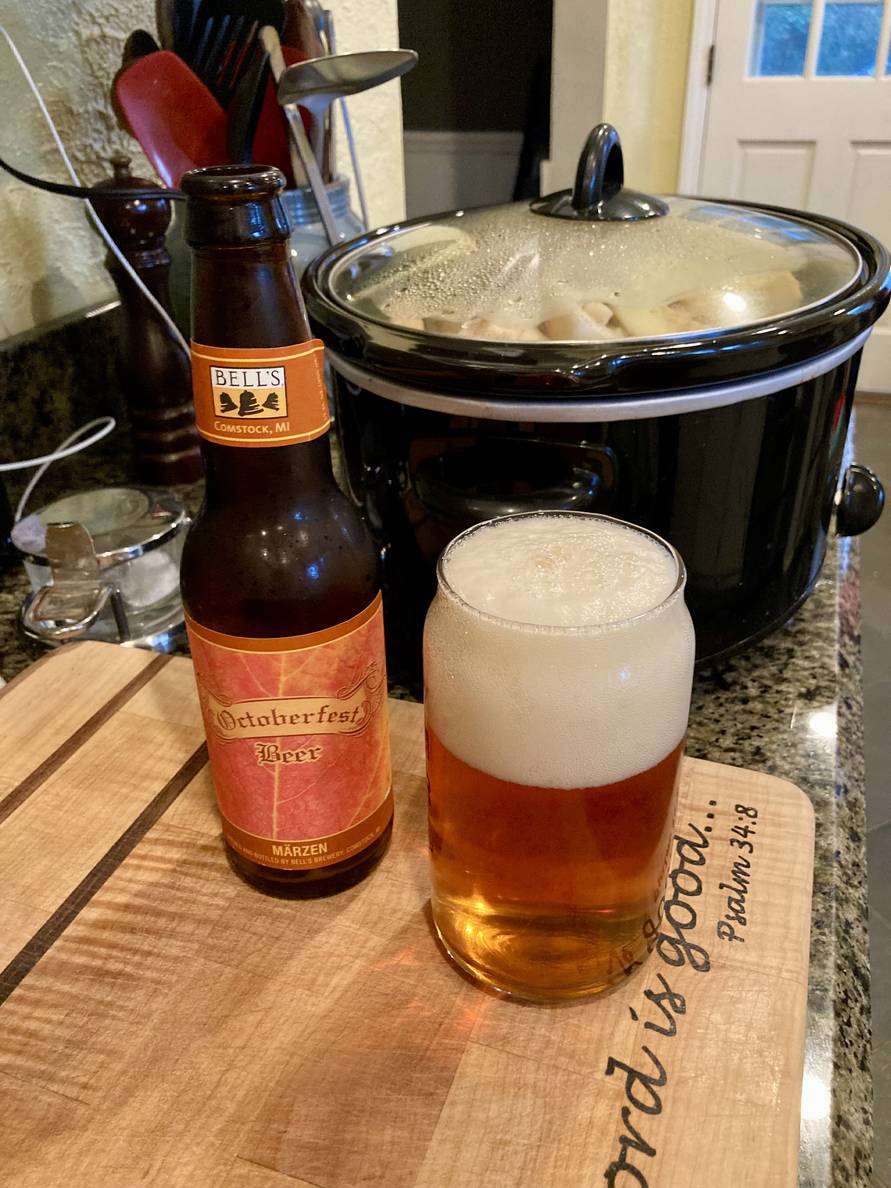A glass of beer and a bottle on a cutting board. The bottle reads Bells, Oktoberfest Beer, Märzen. Behind the beer is a crockpot with apples.