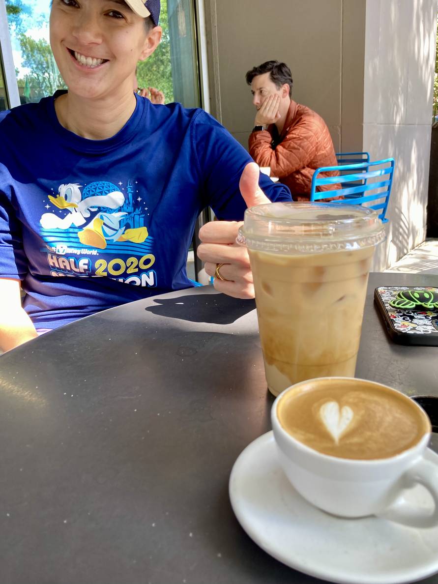 A macchiato and an iced latte outside on a metal table. A woman is smiling and giving a thumbs up on the other side of the table.