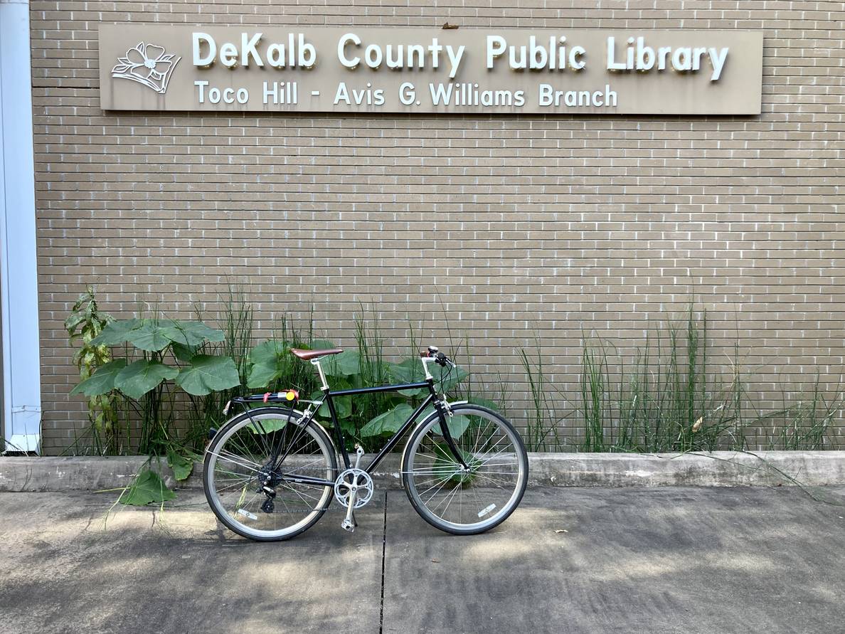 A bicycle in front of a brick wall with a sign saying “DeKalb County Public Library Toco Hill - Avis G. Williams Branch”.
