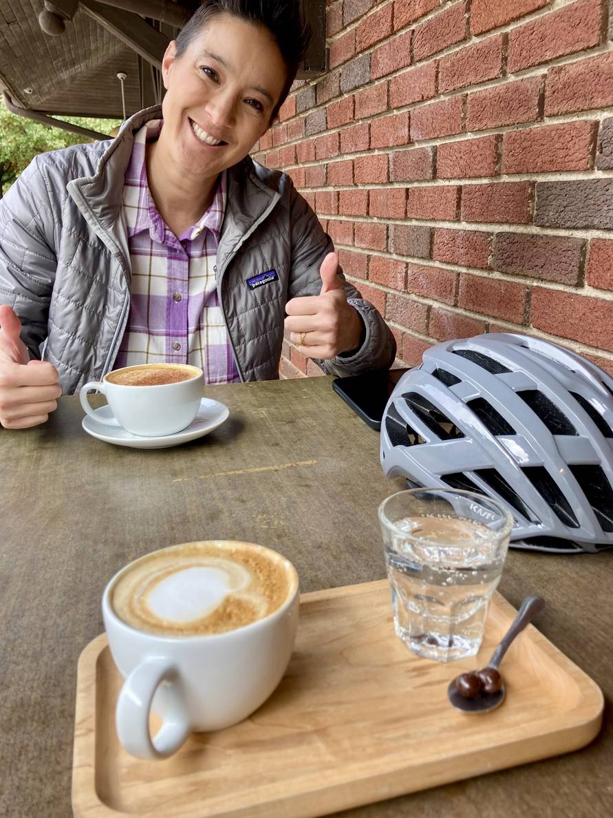 A cappuccino sits on a wooden serving board with a small glass of seltzer. Also on the table is a bike helmet and another coffee drink. On the other side of the table a woman is smiling and giving a thumbs up.