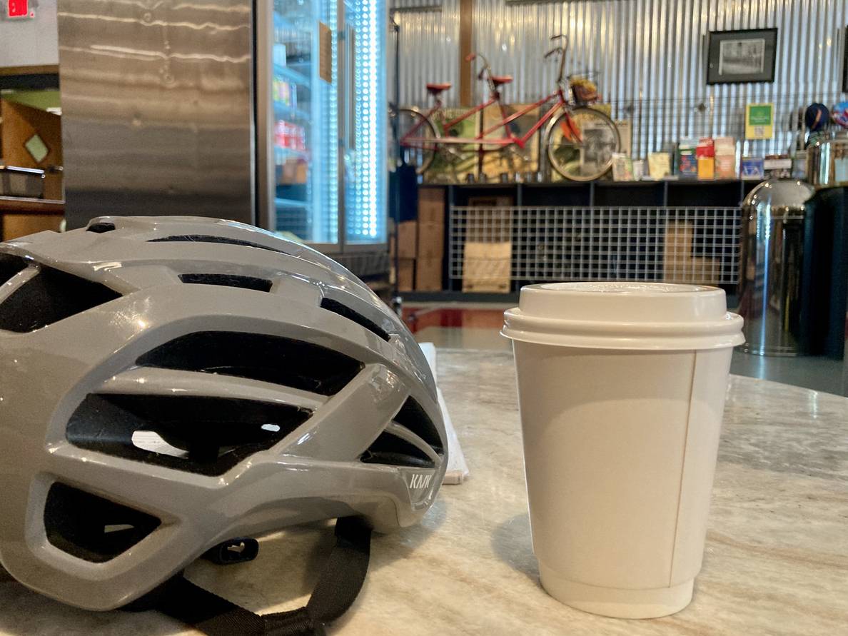 A to-go coffee cup on a table next to a bicycle helmet. A tandem bicycle is on display on the wall in the background.