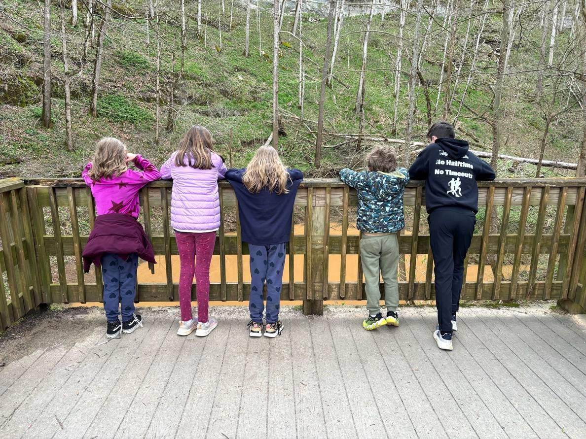Five kids stand at a wood rail. A muddy pond is below them.