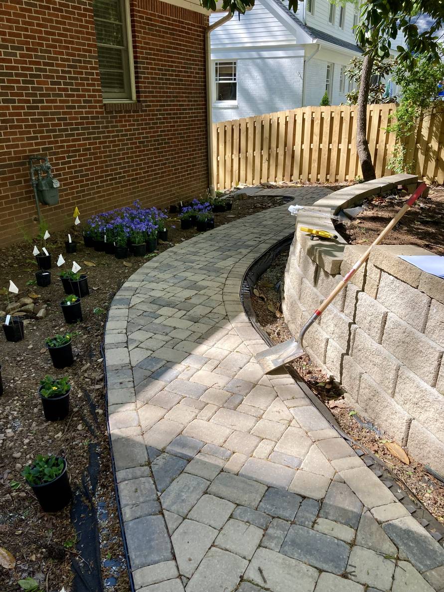 A paver stone path with a garden bed and house to the left, a narrow planting strip and retaining wall to the right. Plants in nursery pots are grouped in the main planting bed.