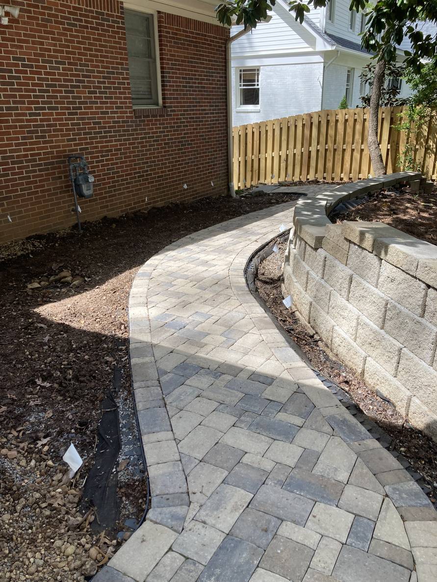 A paver stone path with a garden bed and house to the left, a narrow planting strip and retaining wall to the right. The planting areas are bare dirt and white flags delineate the border.