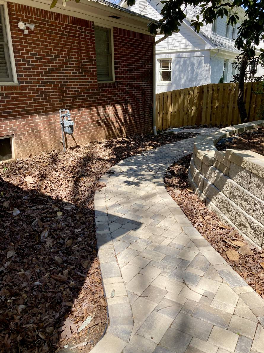 A paver stone path with a garden bed and house to the left, a narrow planting strip and retaining wall to the right. The planting areas are full of leaves.