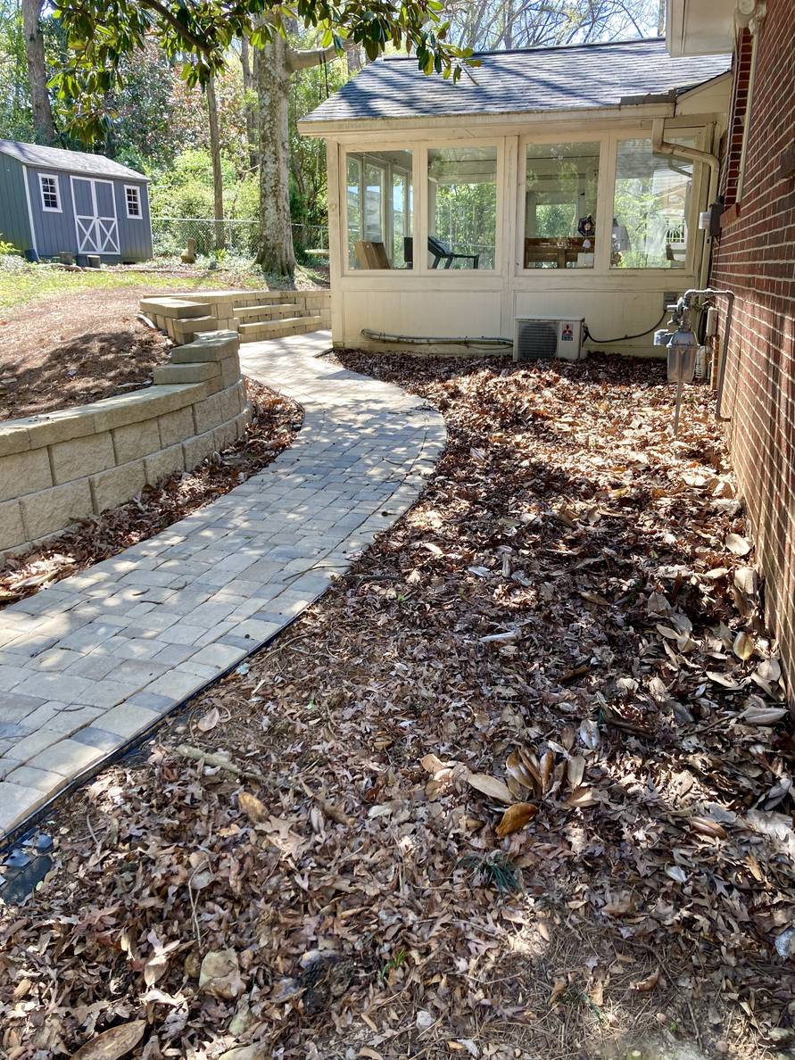 A paver stone path with a garden bed and house to the right, a narrow planting strip and retaining wall to the left. The path winds around a sun porch. The planting areas are full of leaves.