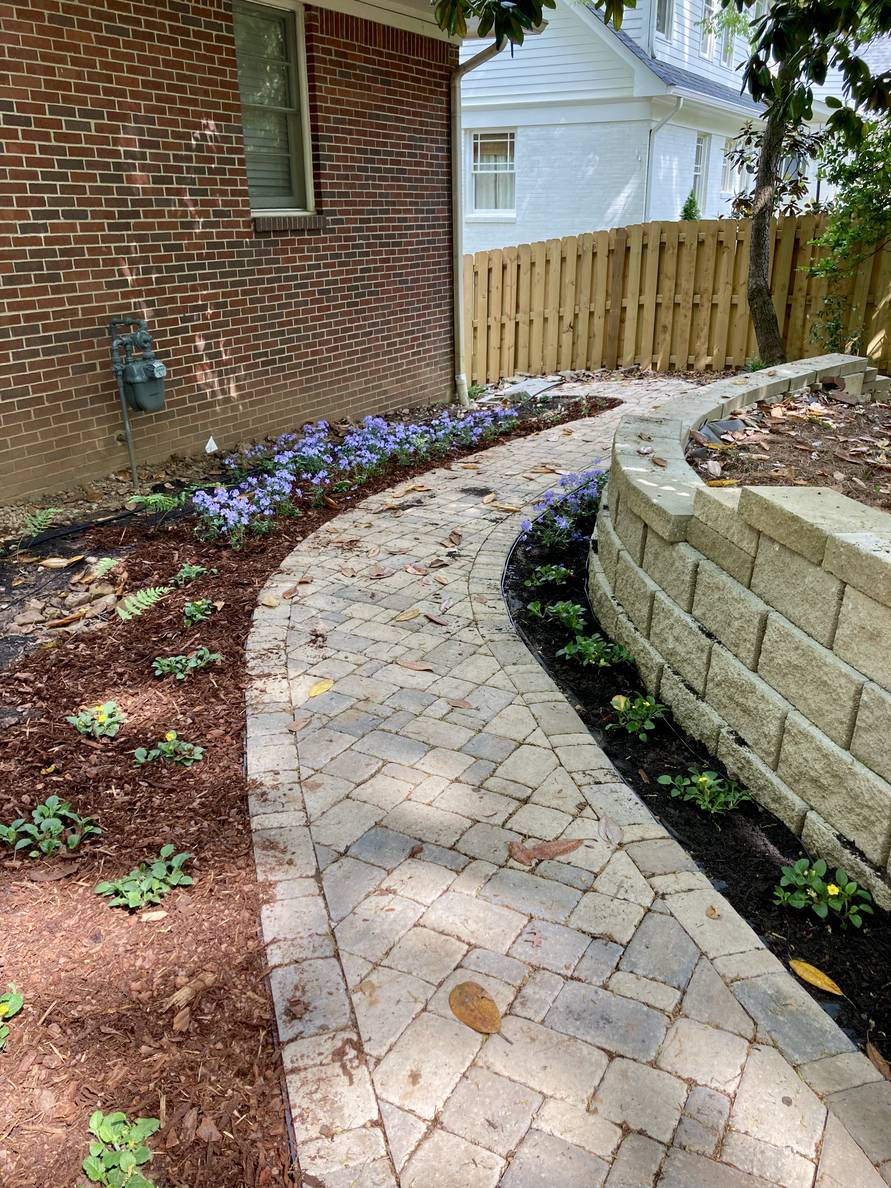 A paver stone path with a garden bed and house to the left, a narrow planting strip and retaining wall to the right. Plants are planted on both sides of the path. Mulch has been partially applied.