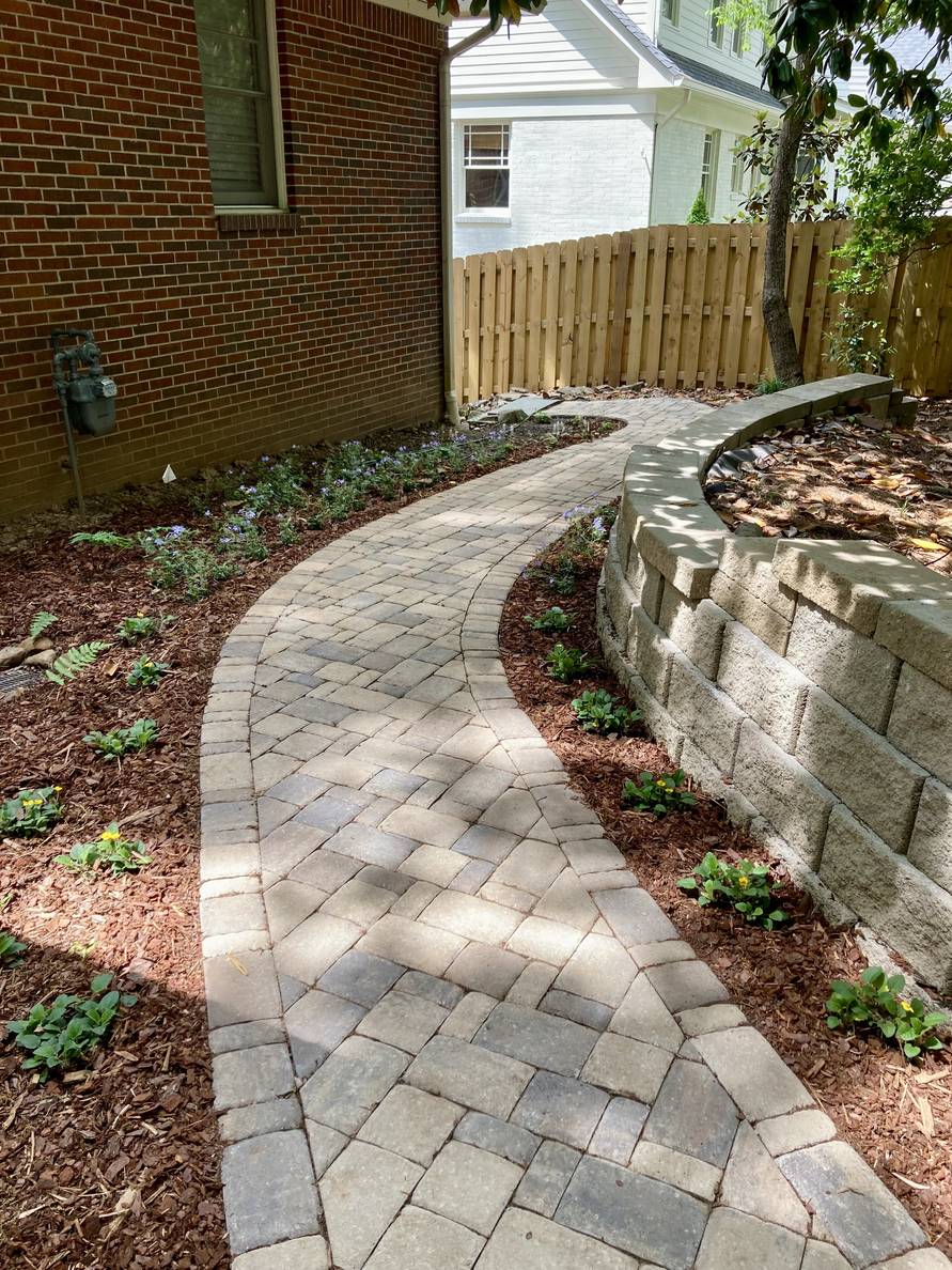 A paver stone path with a garden bed and house to the left, a narrow planting strip and retaining wall to the right. Plants are planted on both sides of the path. The right side and most of the left side has been mulched.