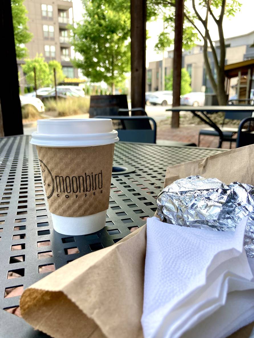 A small to-go coffee cup on an outdoor table next to a foil-wrapped burrito on a paper bag.
