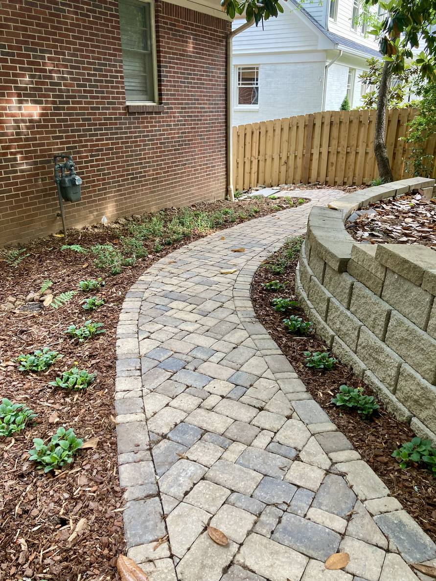 A paver stone path with a garden bed and house to the left, a narrow planting strip and retaining wall to the right. Plants are planted on both sides of the path. Both sides have been mulched.