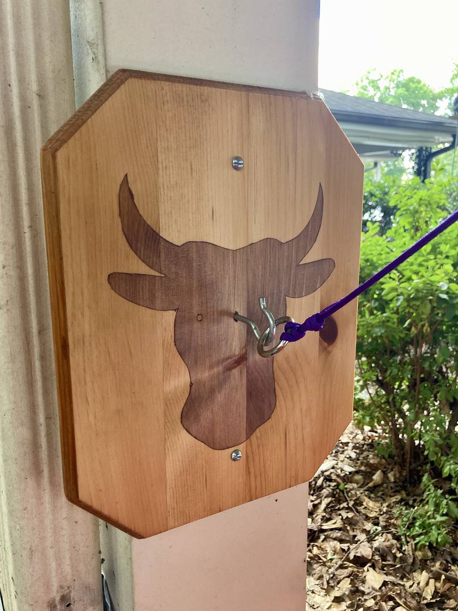 A wooden panel with a stained silhouette of a bull head is mounted to a square column outside. In the middle is a metal hook, on which hangs a metal ring with cord tied to it.
