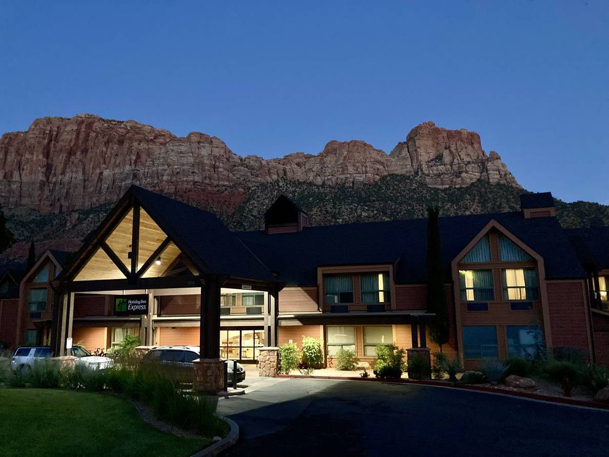 At dusk, a lit Holiday Inn Express is dwarfed by giant desert cliffs behind it.