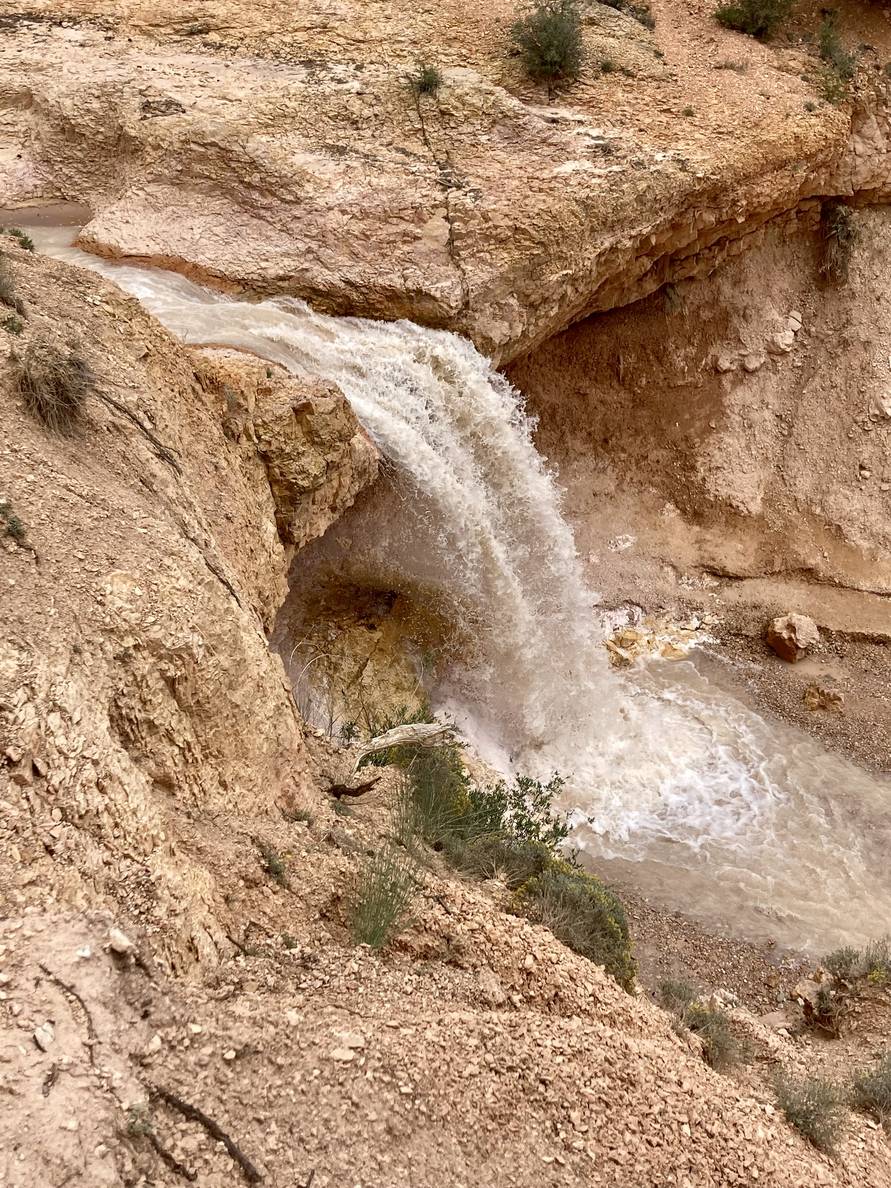 A cloudy waterfall plunges about 10 feet through an arid landscape.