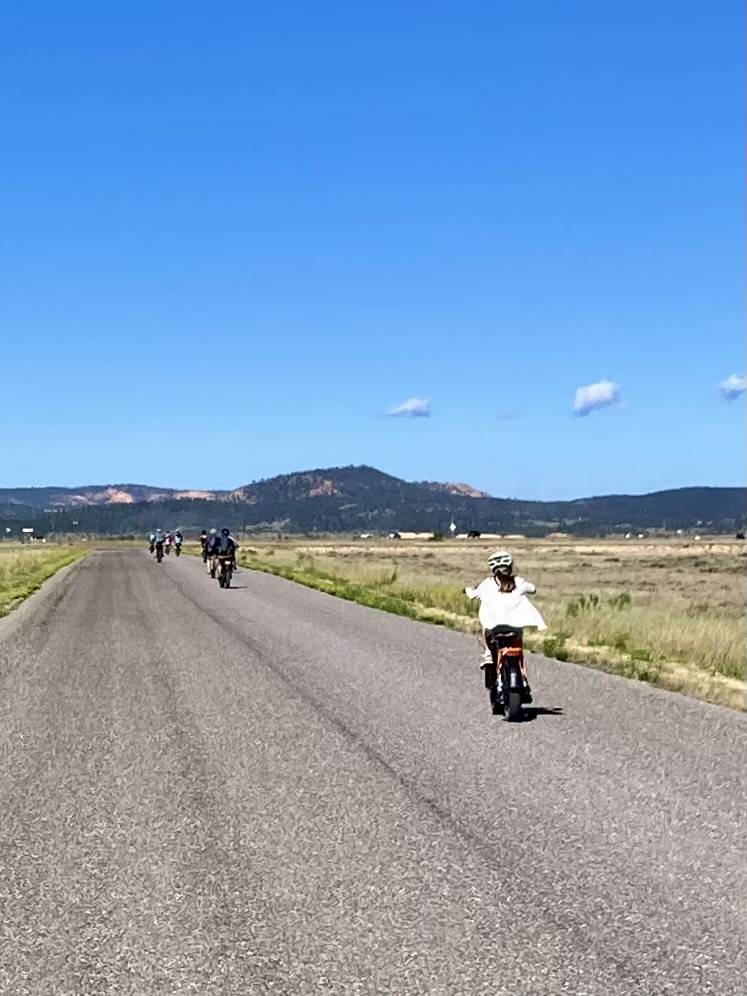 A group on bicycles on a road stretching into the distance. The sky is wide and punctuated with clouds. Some mountains rise in the distance.