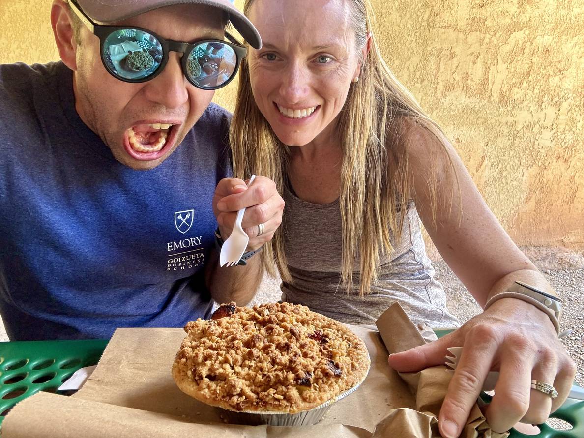 A man and woman sit at a table with a pie before them. The man holds a fork and his mouth is open towards the pie. The woman smiles at the camera.