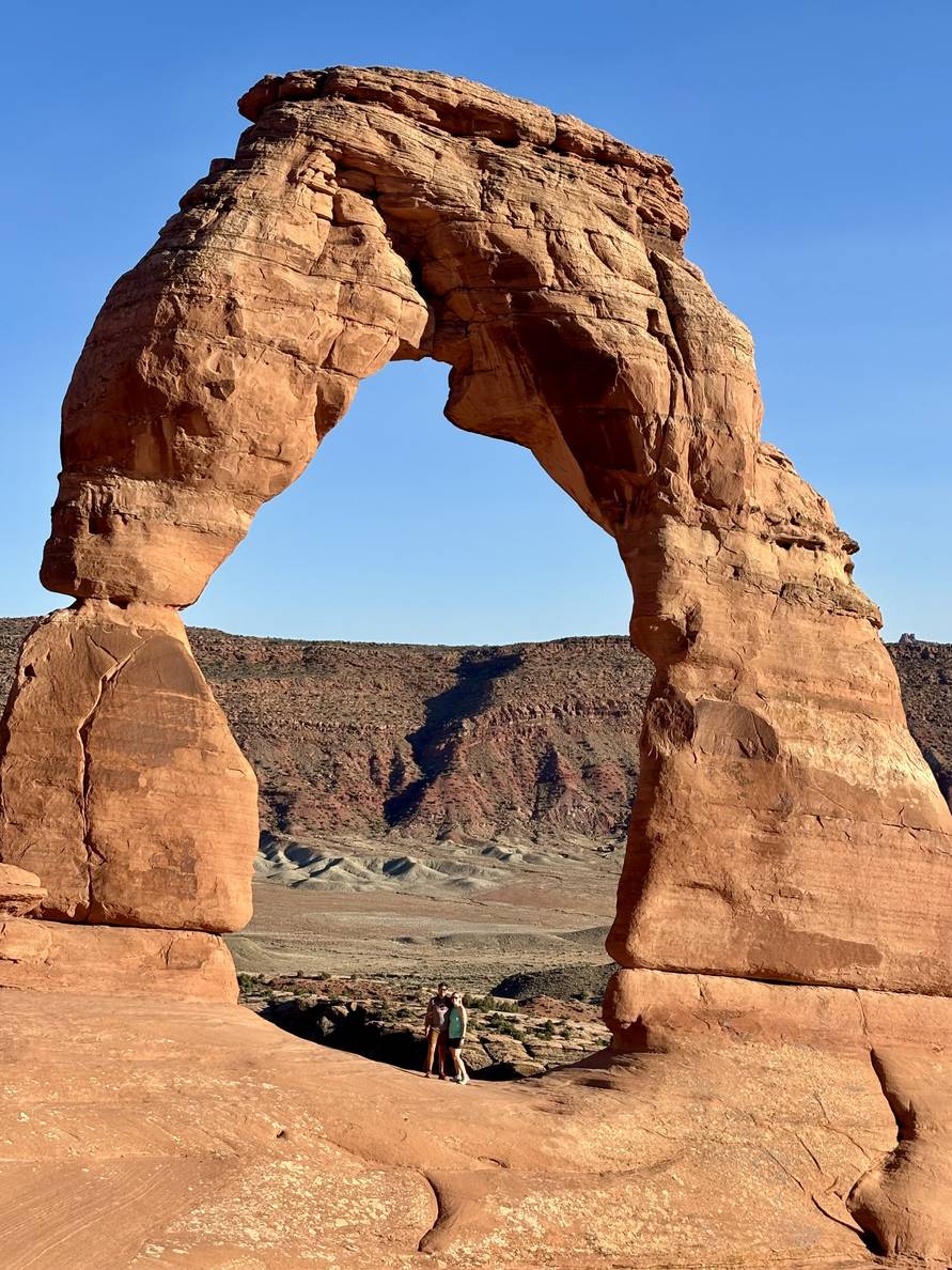 A man and woman are barely visible underneath a red stone arch. Mountains are in the distance.