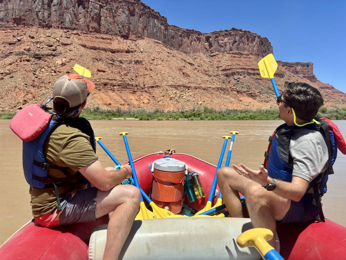 Two men sit in the front of a river raft, on a river, paddles extended towards red rock bluffs rising along the river.