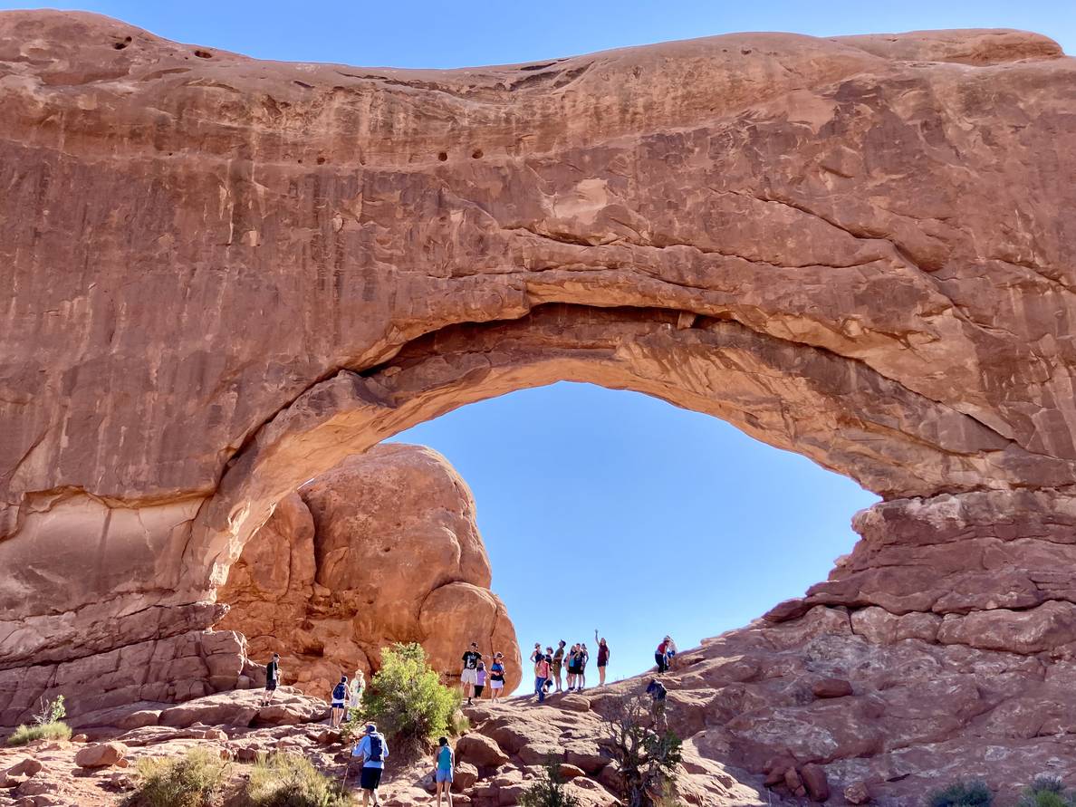 A large stone arch rises against the sky. People, diminutive in comparison are at the bottom of the arch.