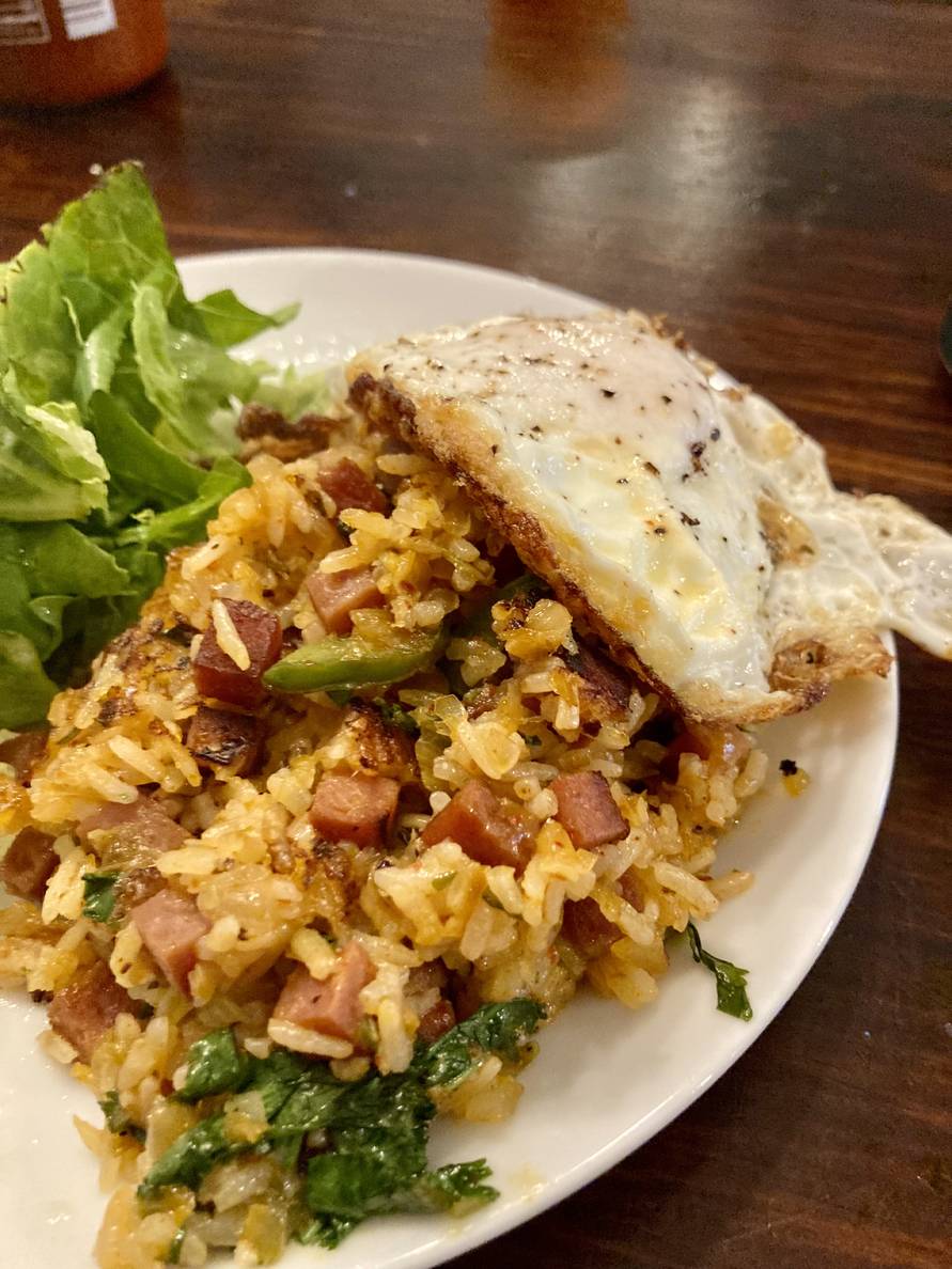 A plate on a table. On the plate is a bit of lettuce and some fried rice with chunks of meat and pieces of cilantro in it. A fried egg is on top of the fried rice.