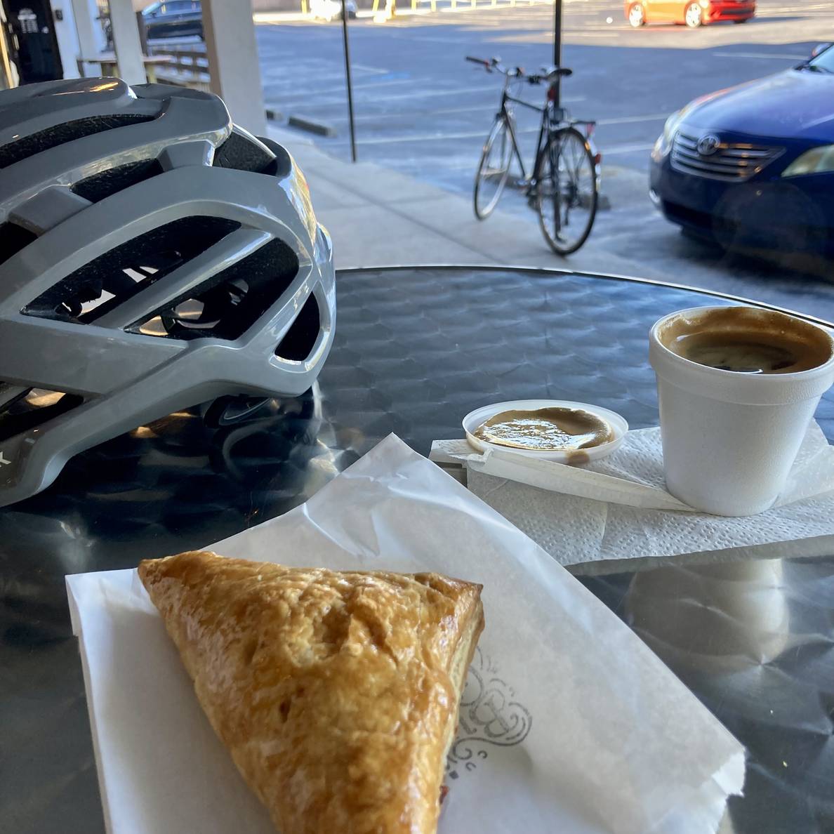 A small cup of Cuban espresso and pastry with bicycle helmet on a metal table. In the background a bicycle is locked to a pole.