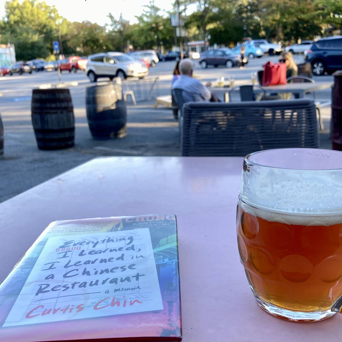 A beer glass on a table with a book titled “Everything I Learned, I Learned in a Chinese Restaurant.” More people, a parking lot, and trees are in the background.