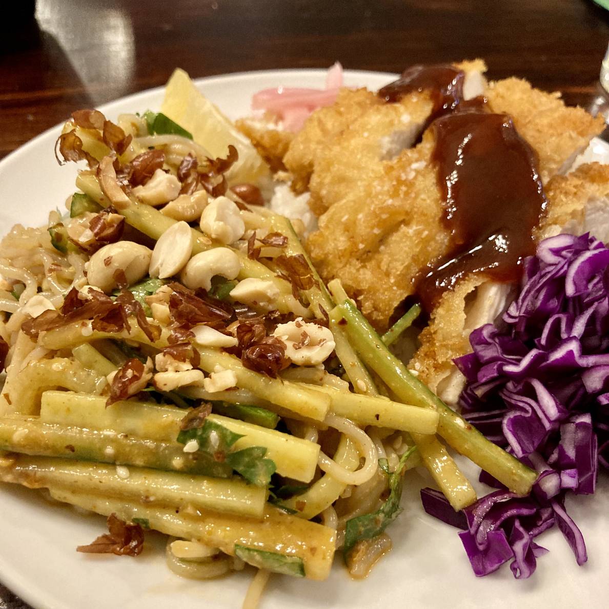 A plate of food with a noodle and cucumber salad, fried chicken, rice, cabbage, pickled onions, and a lemon slice.