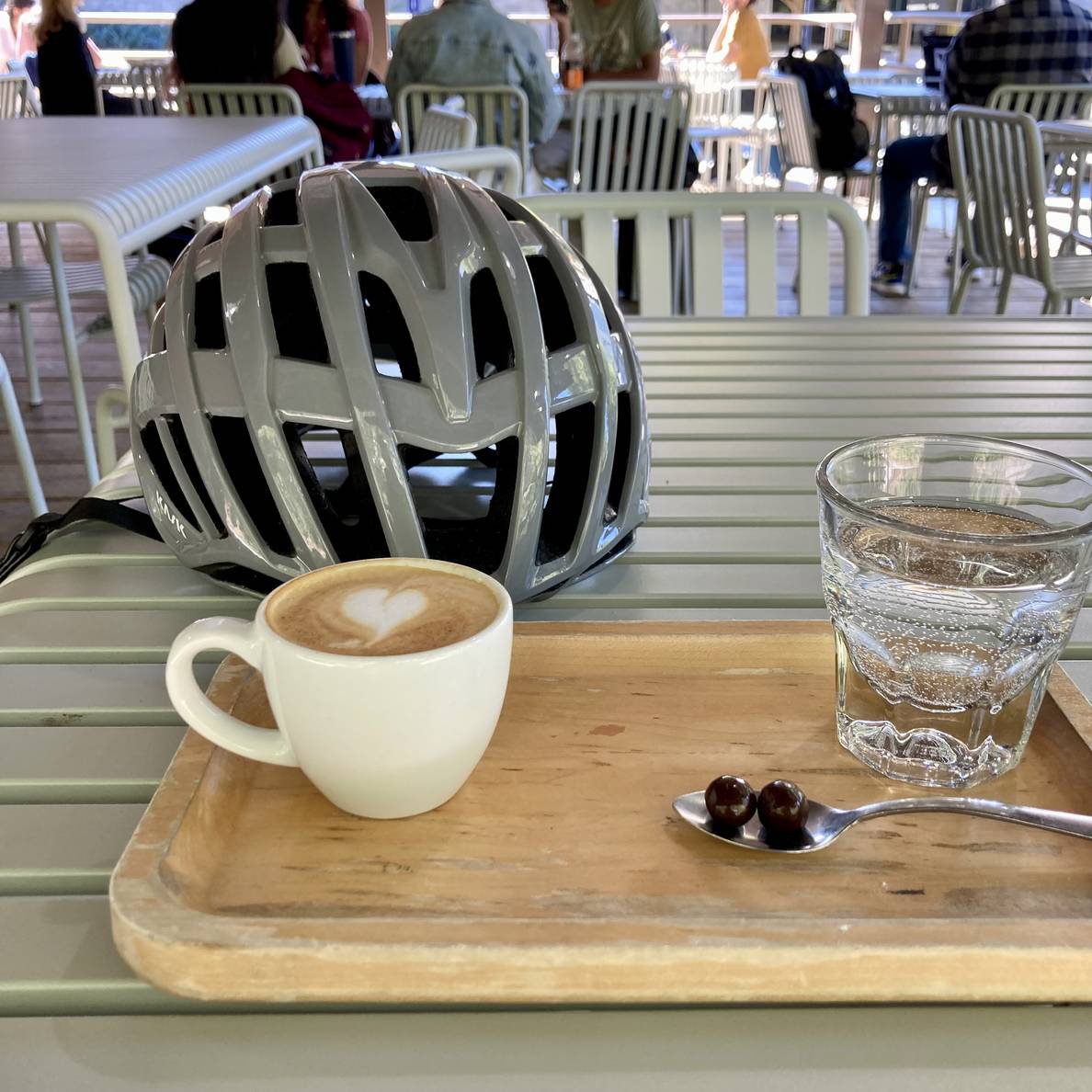 An espresso cup and glass of water on a wooden tray outside on a table with a bicycle helmet.