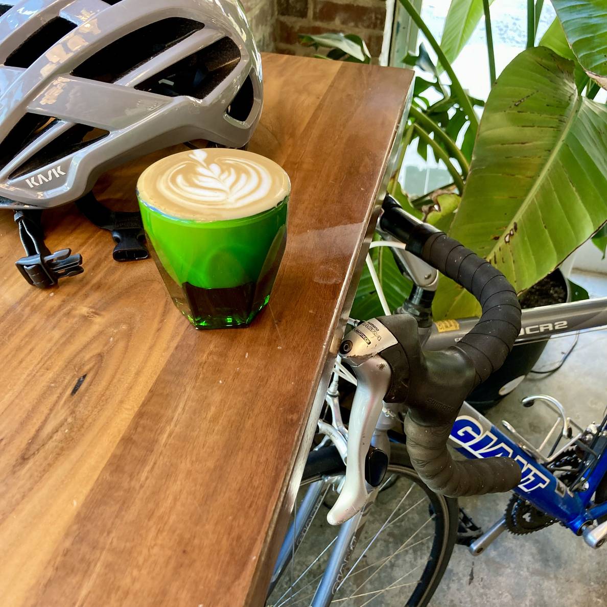 An espresso drink with latte art on wooden counter with a bicycle helmet. Below the counter is the front of a bicycle.