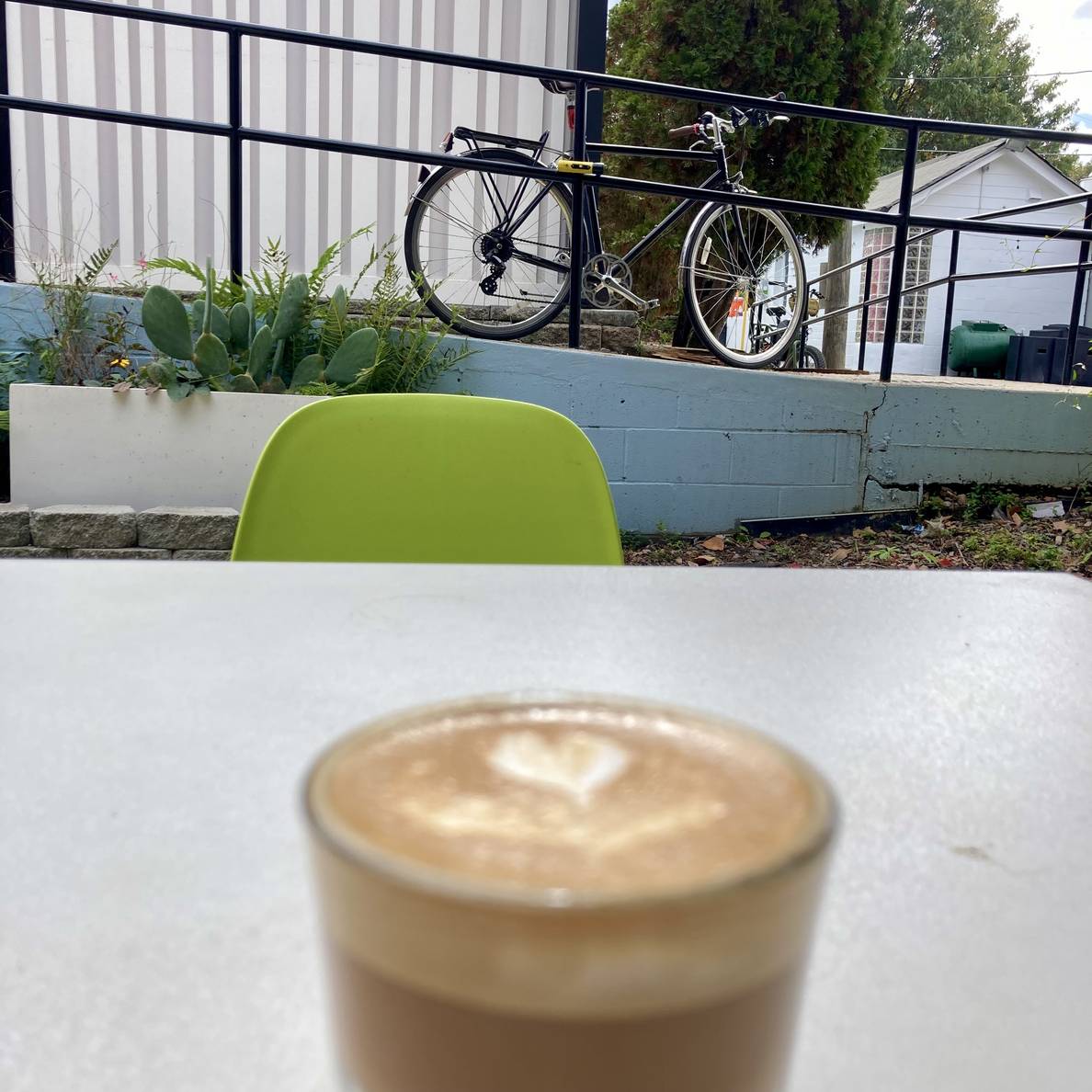 In the foreground, but out of focus, is a cortado on an outside table. In the background, but in focus, is a bicycle locked to a railing.