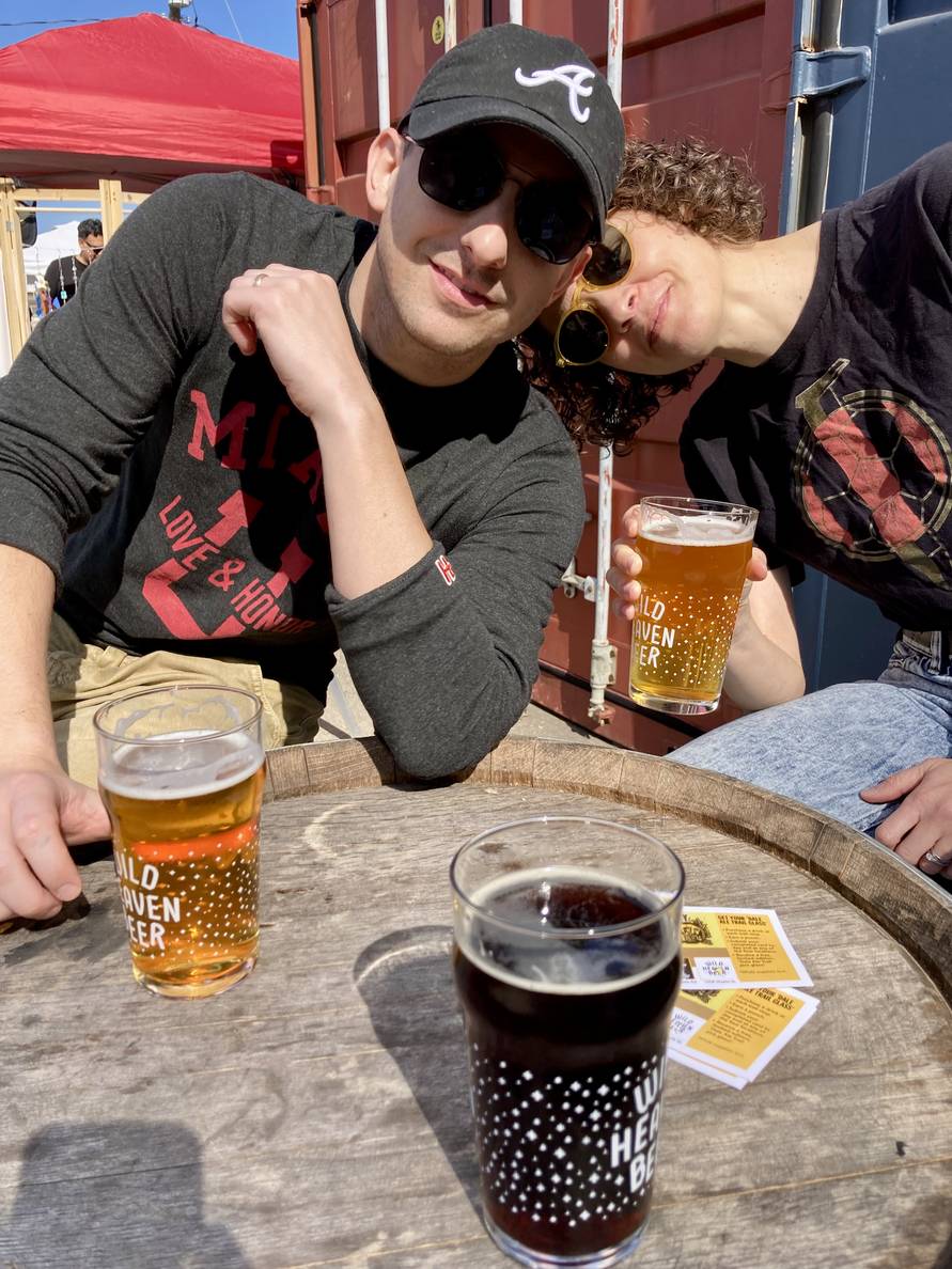 Two people sitting behind a wooden barrel table. One is holding a beer. Two more beers are on the table.