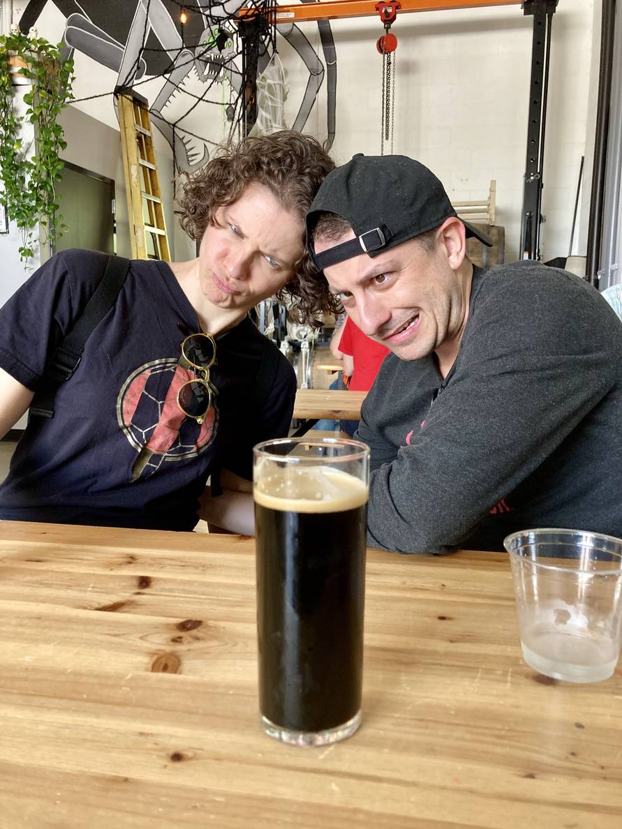 Two people behind a wooden table with funny expressions. A beer is on the table.