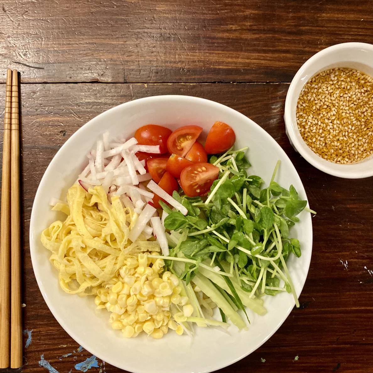 An array of vegetables top a bowl on a wooden table. Next to the bowl are chopsticks and a small bowl of sauce.